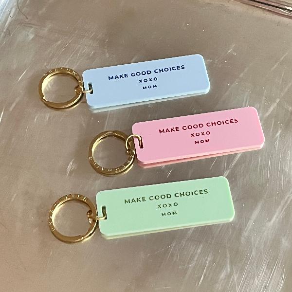 Three keychains with the inscription 'Make Good Choices' in different color schemes: light blue with navy engraving, pale pink with hot pink, and light green with dark green engraving. Each keychain has a gold-toned keyring and the text 'XOXO, Mom'.
