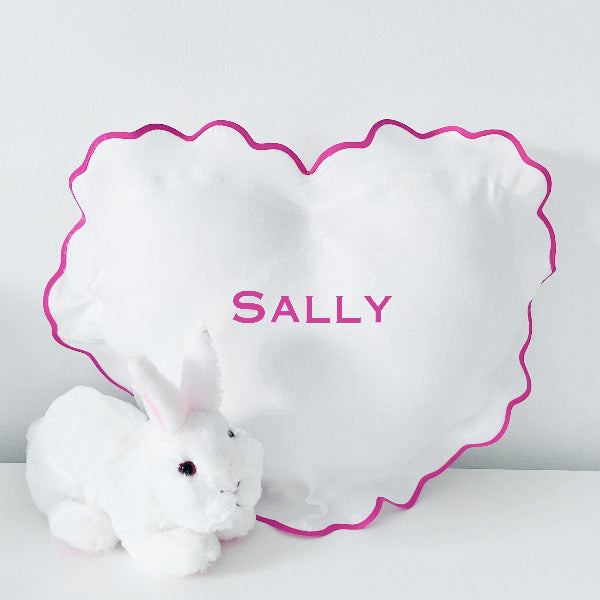 A heart-shaped white linen baby pillow with a scalloped edge and hot pink trim. The pillow has the name 'Sally' monogrammed in pink lettering.