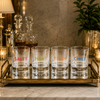 Four clear lowball glasses with colorful text reading 'SALTY', 'NEAT', 'FIZZY', and 'CHILL' in pink, green, orange, and blue, arranged on a gold-toned tray in a warmly lit bar setting.
