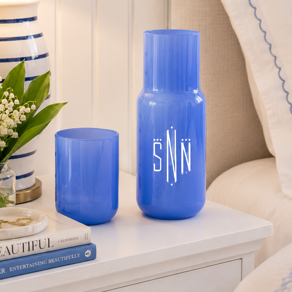 Two blue glass vases, one tall and one short, displayed on a white bedside table next to a striped lamp, flowers, and a bed with white pillows.