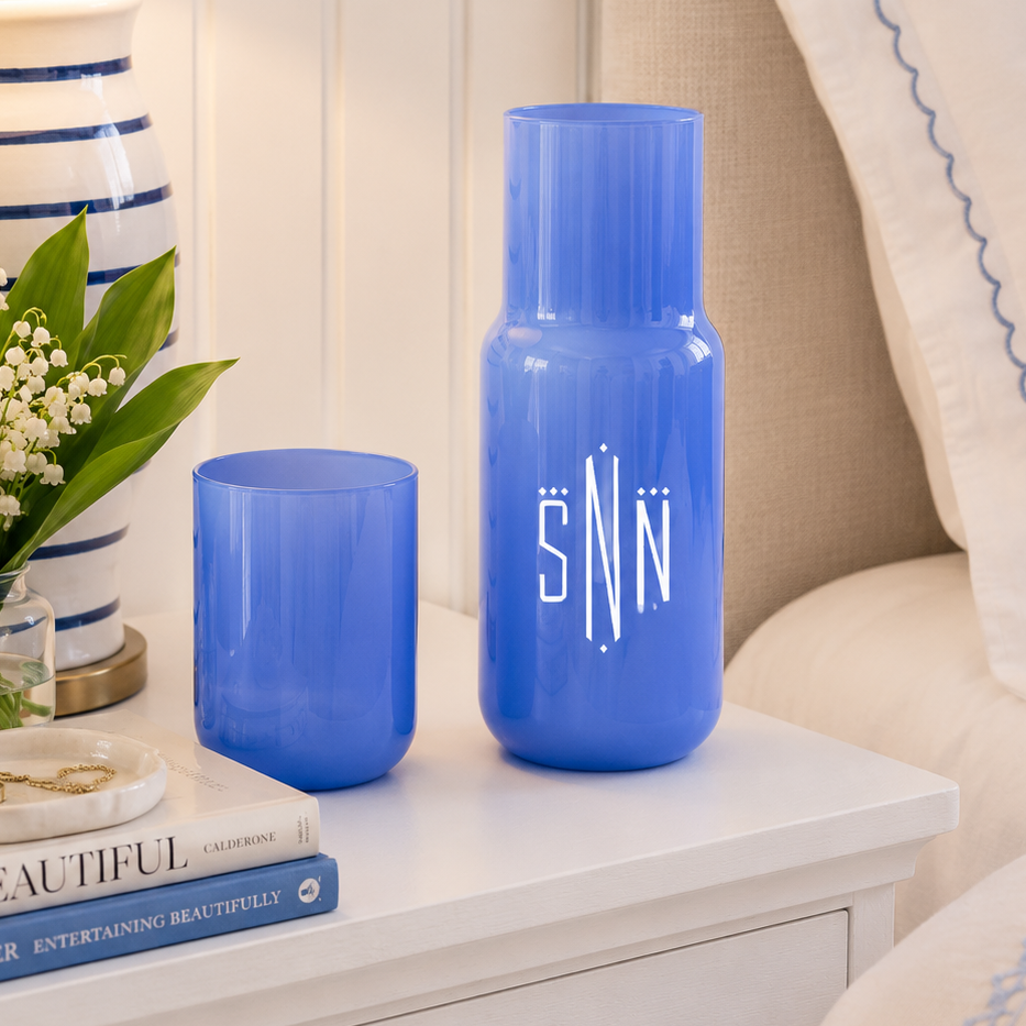 Two blue glass vases, one tall and one short, displayed on a white bedside table next to a striped lamp, flowers, and a bed with white pillows.