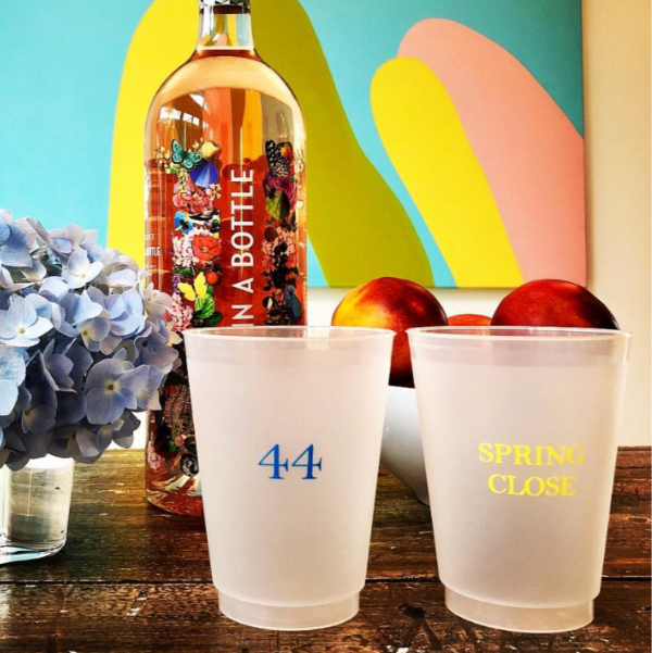 Bottle of alcohol with decorative bottle cap, two cups labeled '44' and 'Spring Close', and flowers on a wooden surface.