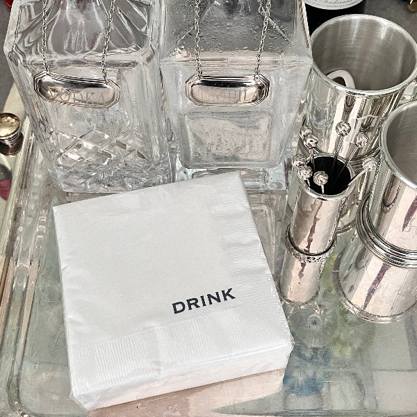 A set of white cocktail napkins with the word 'DRINK' printed on them, displayed on a table with glassware and metalware in the background.