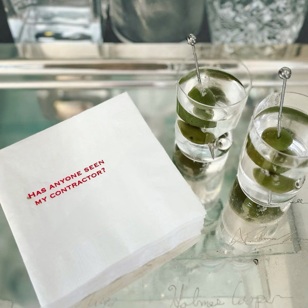 A set of paper napkins with the text 'HAS ANYONE SEEN MY CONTRACTOR?' printed on them, displayed on a table with two glasses of martini.