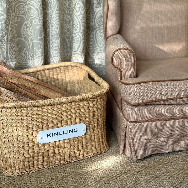 Wicker basket labeled 'Kindling' next to a beige armchair with patterned curtains.