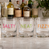 Four clear lowball glasses with the words SALTY, NEAT, FIZZY, and CHILL printed in pink, green, orange, and blue respectively, arranged on a light marble surface with blurred bottles and decor in the background.