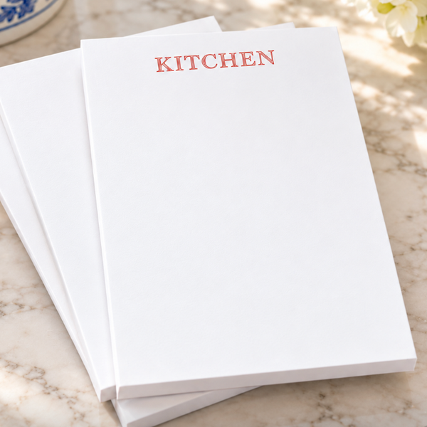 Three white rectangular cards or sheets stacked on a marble surface, with the top sheet printed in red text reading “KITCHEN.” A blue-and-white ceramic object is partially visible in the top left, and flowers appear in the top right.