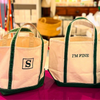 Two tote bags with green straps on a purple surface, one with a 'S' logo and the other with 'I'M FINE' text.