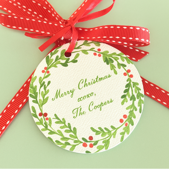 Round Christmas gift tag with green wreath design, red berries, and red ribbon on a light green background.