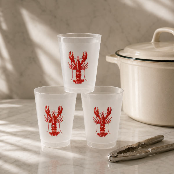 Clear glasses with red lobster design on a marble surface with a white pot and tongs.