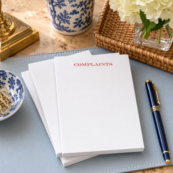 A stack of white notepads with the word 'COMPLAINTS' printed in red at the top, styled on a light blue leather pad with a navy and gold pen nearby, alongside decorative blue-and-white ceramic dishes, flowers, and a wicker tray.