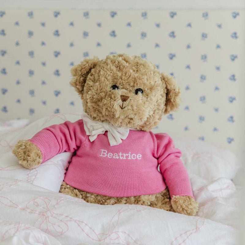 Teddy bear wearing a pink sweater with 'Beatrice' on it, sitting on a textured surface.