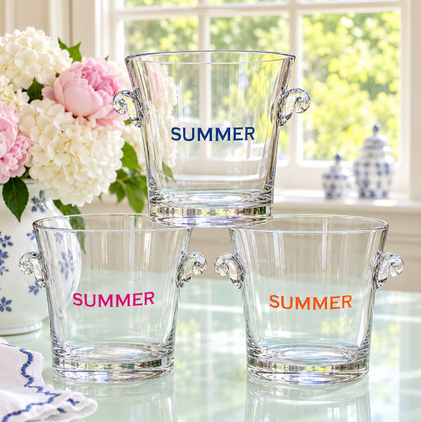 Three clear glass bucket-style tumblers with side handles are stacked on a reflective table in a bright room. The top bucket is centered behind the two front buckets, and each cup is printed with the word "SUMMER" in a different color: blue on the top bucket, pink on the left front bucket, and orange on the right front bucket.