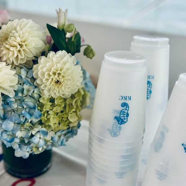 Stacks of white disposable cups with blue text and a logo next to a floral arrangement.