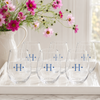 Six clear stemless wine glasses with blue monogrammed initials 'H' on a white tray, styled in a bright kitchen setting with a white pitcher of pink flowers in the background.
