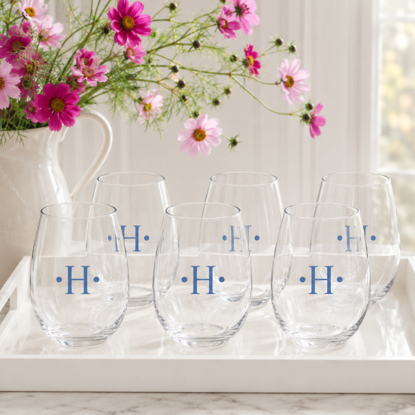 Six clear stemless wine glasses with blue monogrammed initials 'H' on a white tray, styled in a bright kitchen setting with a white pitcher of pink flowers in the background.