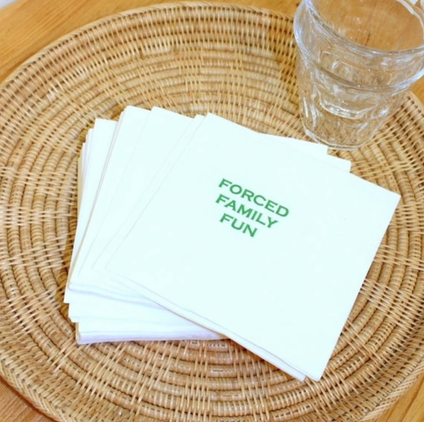 Stack of white napkins with 'FORCED FAMILY FUN' text on a woven placemat.