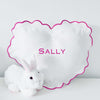A heart-shaped white linen baby pillow with a scalloped edge and hot pink trim. The pillow has the name 'Sally' monogrammed in pink lettering.
