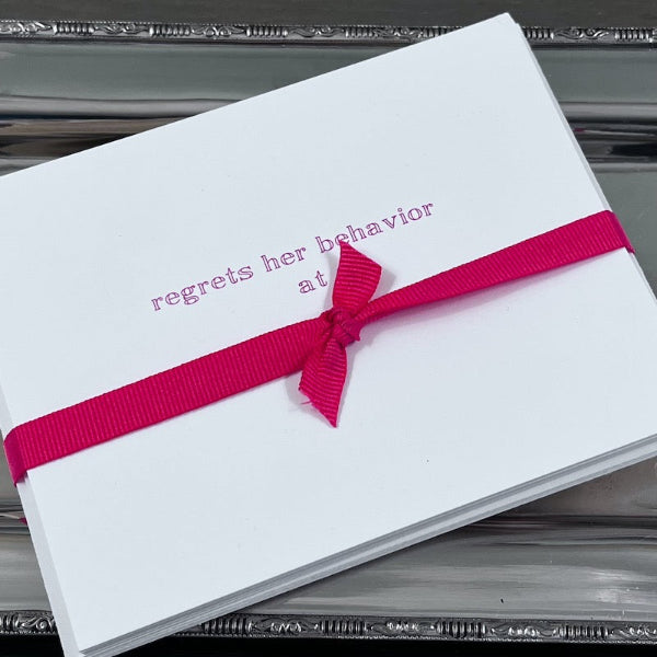 A set of notecards with the text 'regrets her behavior' on the front, sealed with a hot pink ribbon.